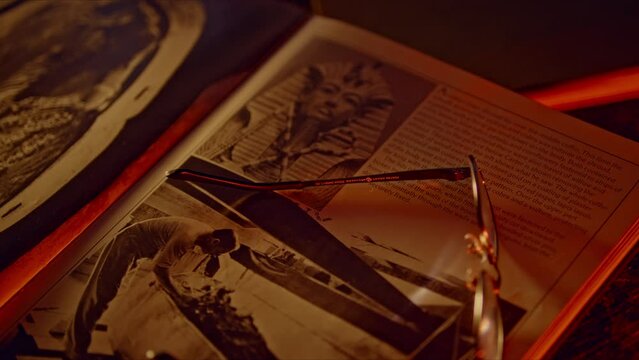 Camera racking focus across rare ancient Egyptian archaeology book. Black and white photo of Tutankamen's death mask and Howard Carter excavation featured. Cinematic lighting.