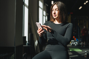 Portrait of resting Young brunette woman in sport active wear using mobile in fitness club gym