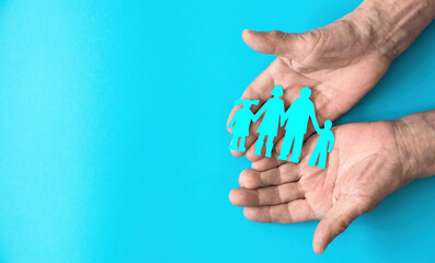 The hands of an elderly person hold a paper-cut silhouette of a family on a blue fog. Copyspace on the left. Family day, family traditions concept. Family tree, succession of generations.