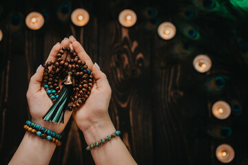 Woman holds in hand wooden mala beads strands used for keeping count during mantra meditations. Weaving and creation. Wooden background with candles and feathers. Spirituality, religion, God concept.