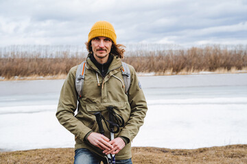 A young handsome guy in a yellow hat looks into the camera, a hipster traveler stands in nature by the lake, cold weather wind blows hair, cloudy sky. © Aleksey