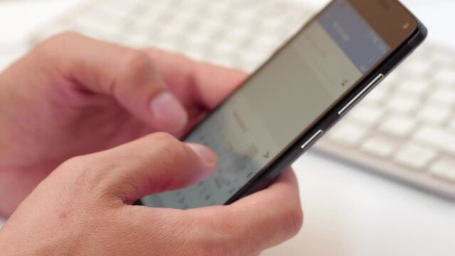A man in an office at a table with a keyboard is typing a text message on a smartphone.