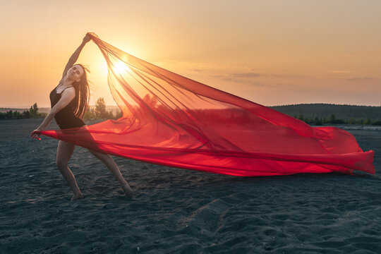 Elegant girl performs dance moves on sand holding up long red fabric against sunset. - Powered by Adobe