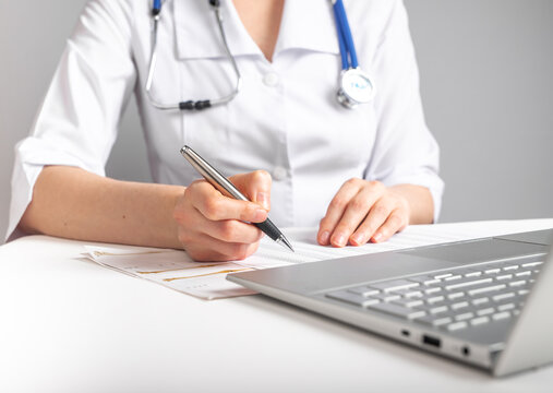 Doctor with stethoscope writing with pen about patient treatment or scheduling appointments. Woman in lab coat sitting at table with laptop. Health care and medicine concept. High quality photo