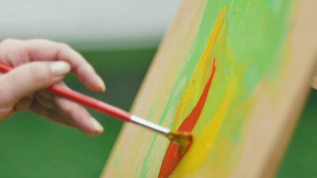 A brush with wide bristles draws a red curl with bright saturated paint on canvas on a wooden easel