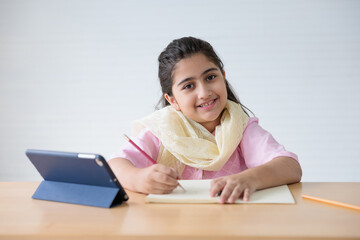 portrait cute Indian girl writing on notebook and online learning class from tablet, education...