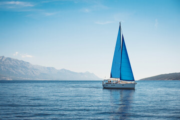 White yacht with blue sails in the sea against a background of blue sky and mountains - obrazy, fototapety, plakaty