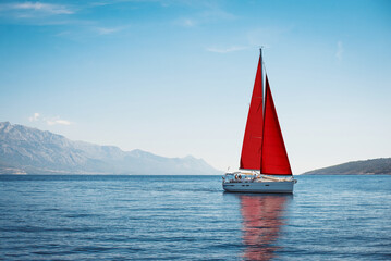 White yacht with red sails in the sea against a background of blue sky and mountains