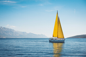 White yacht with yellow sails in the sea against a background of blue sky and mountains