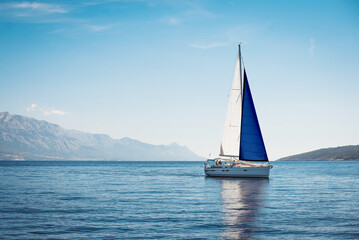 A white yacht with sails the color of the Finnish flag in the sea against a background of blue sky and mountains
