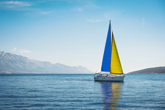 A white yacht with sails the color of the Ukrainian flag in the sea against a background of blue sky and mountains