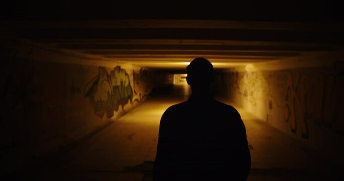 Silhouette of the back of a man walking in a dark underpass. Dark scary movie scene at night city. Handheld camera shot
