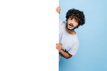 Excited indian or arabian confused curly guy, peeking out from behind white vertical advertisement...