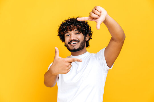 Joyful happy positive indian or arabian millennial guy in basic white t-shirt, making imaginary frame cadre of camera taking shot, standing on isolated orange background, looks at camera, smiling
