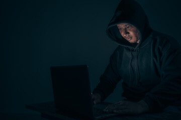 A hacker wears a long-sleeved shirt with a hood covering his head. Sitting in a dark room using a laptop The concept of identity theft on the Internet camp network. with copy space.