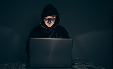 A hacker wears a long-sleeved shirt with a hood covering his head. He was sitting in a dark room using his laptop and had a lot of dollars on the table. Data theft concept. with copy space.