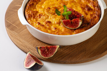 curd casserole with pumpkin in a round white shape on a wooden board for serving on a white table decorated with figs