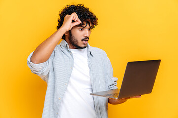 Puzzled shocked confused Indian or Arabian guy, in casual wear, take off glasses, holds an open laptop in hand, looks surprised at it, while standing on isolated orange background, get unexpected news