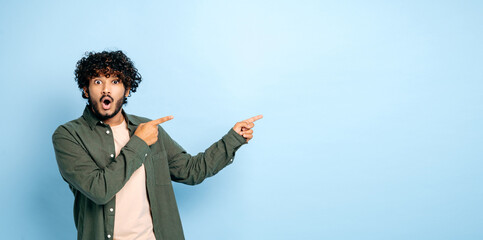 Panoramic photo of amazed shocked indian or arabian guy in casual wear, points fingers to the side at mock-up empty space, stands on isolated blue background, looks surprised at the camera, astonished