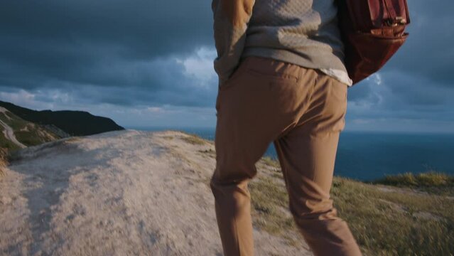 Close-up pano shot of young male hipster traveler walk with a bag to the edge of cliff vith stunning view in front of beautiful sky sunset and seascape. Traveling alone. Discover new horizons.