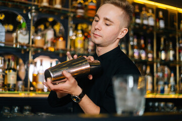 Close-up of bartender in black clothes mixing ingredients of alcoholic cocktail by shaking shaker standing behind bar counter, on blurred background of shelves with alcoholic drinks, selective focus.