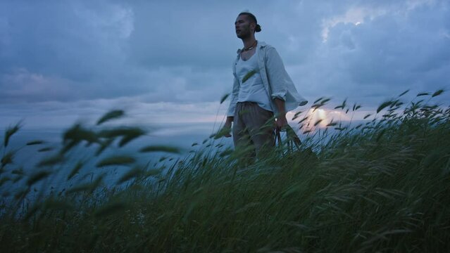 Brave young attractive handsome stranger traveler man stands and looks at beautiful wheat field in windy moody north mountain landscape with bag on his shoulder. Cinematic freedom people concept