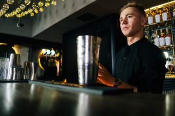 Close-up low-angle view of confident barman making refreshing alcoholic cocktail standing behind bar counter in modern dark nightclub, on background of shelves with different alcoholic drinks.