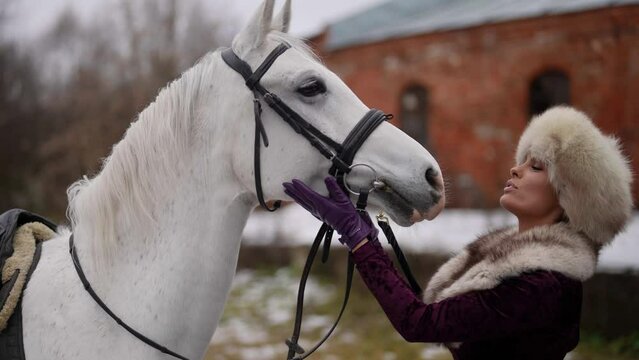 beautiful woman in fur hat and coat is talking with white stallion in horse yard, medieval scene