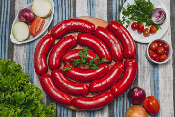 Linguiça calabresa vermelha grande crua em gomos sobre tábua de madeira em toalha azul e cinza acompanhado de tomate cereja cebola roxa alface salsa in natura cortados