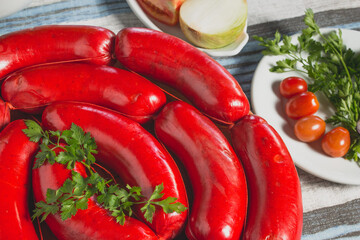 Linguiça calabresa vermelha grande crua em gomos sobre tábua de madeira em toalha azul e cinza acompanhado de tomate cereja cebola roxa alface salsa in natura cortados