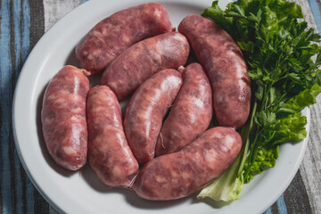 Linguiça calabresa mista porco crua em gomos grandes em prato branco sobre toalha azul e cinza acompanhado de salsa in natura