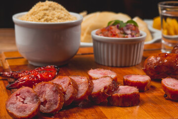 Churrasco de linguiça calabresa inteira com pedaços cortados no entorno e acompanhado de pão mandioca frita vinagrete e farofa sobre tábua de carne em mesa de bar com copo de cerveja e pimenta