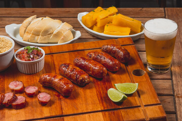 Churrasco de linguiça calabresa inteira com pedaços cortados no entorno e acompanhado de pão mandioca frita vinagrete e farofa sobre tábua de carne em mesa de bar com copo de cerveja pedaços de limão