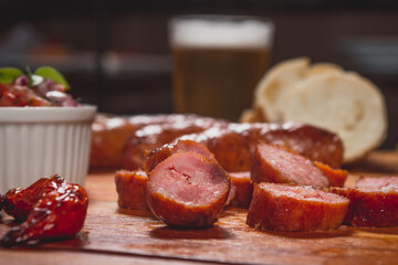 Churrasco de linguiça calabresa inteira com pedaços cortados no entorno e acompanhado de pão mandioca frita vinagrete e farofa sobre tábua de carne em mesa de bar com copo de cerveja e pimenta