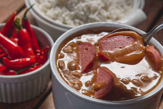 Feijoada com feij&atilde;o carioca em tigela branca servido com concha metal com lingui&ccedil;a calabresa crua acompanhada de arroz branco pimenta sobre mesa de bar em madeira