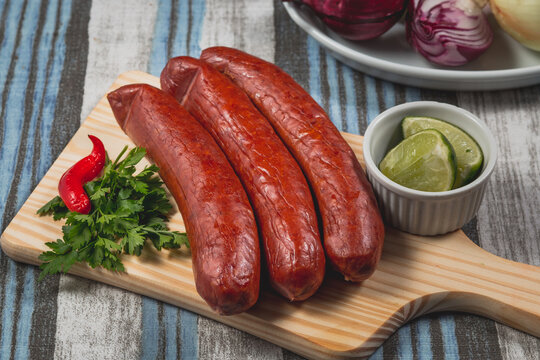 Lingui&ccedil;a calabresa grande crua em gomos sobre t&aacute;bua de madeira em toalha azul e cinza acompanhado de cebola roxa alface salsa lim&atilde;o pimenta in natura cortados em potes pequenos