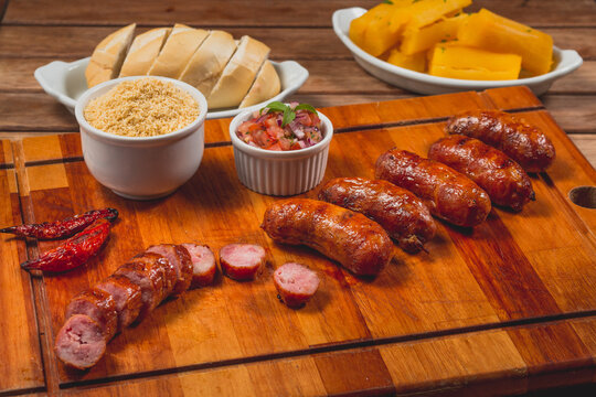 Churrasco de lingui&ccedil;a calabresa inteira com peda&ccedil;os cortados no entorno e acompanhado de p&atilde;o mandioca frita vinagrete e farofa sobre t&aacute;bua de carne em mesa de bar com copo de cerveja e pimenta