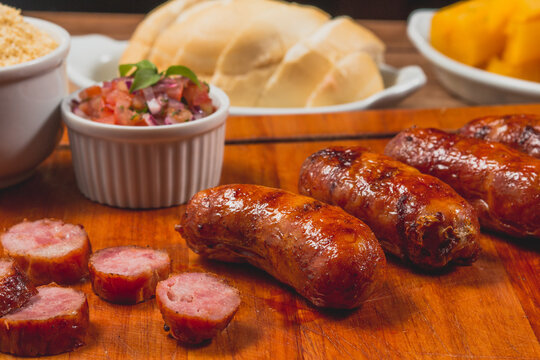 Churrasco de lingui&ccedil;a calabresa inteira com peda&ccedil;os cortados no entorno e acompanhado de p&atilde;o mandioca frita vinagrete e farofa sobre t&aacute;bua de carne em mesa de bar com copo de cerveja peda&ccedil;os de lim&atilde;o