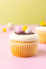 Close up Easter vanilla cupcake with cream cheese, colorful candy eggs, chocolate sprinkles on a pink and yellow background.