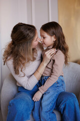 Lovely family mother and daughter embracing and kissing each other indoors. Happy sweet woman with little 6-year-old girl in casual clothers: turtlenecks and jeans sitting on sofa, hugging and sm