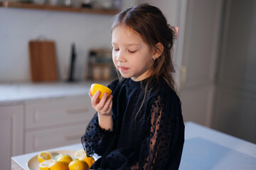 Obraz premium Little baby girl trying and showing lemons in kitchen at home. Child in black holiday dress playng and joking with frits. Happiness and joy cooking and living healthy life