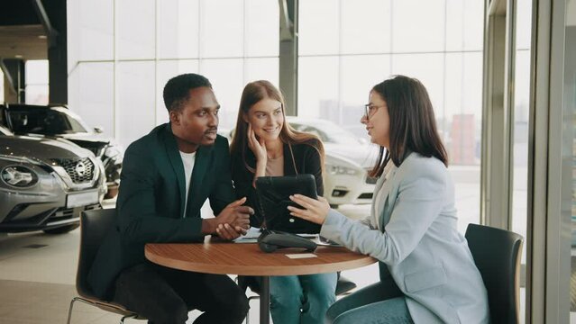 Caucasian saleswoman using digital tablet while working with multi ethnic couple at car salon. Happy man and woman talking with competent manager while buying auto. Talking with dealer at auto salon.