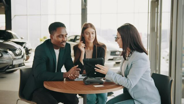 African man and caucasian woman talking with female car dealer at auto salon while making expensive purchase. Couple choosing new vehicle together. Saleswoman using tablet while working with clients.