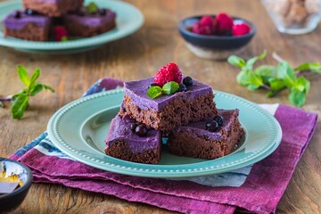 Dessert, sliced chocolate brownies with a layer of ricotta or cream cheese and blueberry puree on a green plate on a brown wooden background. American cuisine.