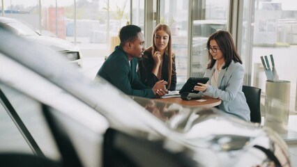 Cheerful multicultural couple sitting with saleswoman at modern salon. Successful agreement of car purchsing. Auto business concept. Manager using digital tablet while working with buyers at showroom - Powered by Adobe