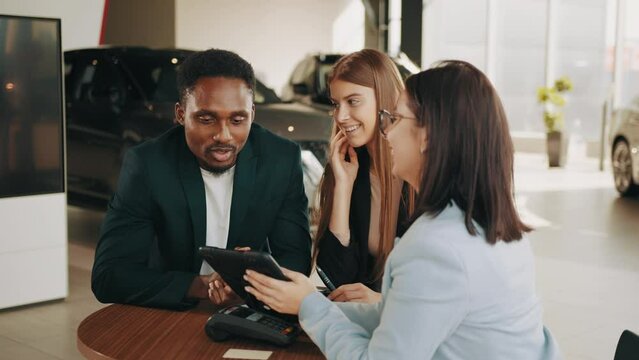 Competent car dealer sitting with multiracial couple at auto salon and making contact about purchasing of new vehicle. Female seller consulting diverse couple about car purchase.