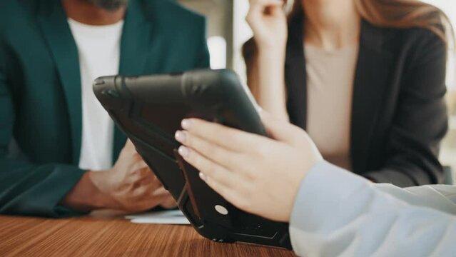 Hands holding tablet computer with blurred background of African man and caucasian woman talking with female car dealer at auto salon. Couple choosing new vehicle together., buying new car