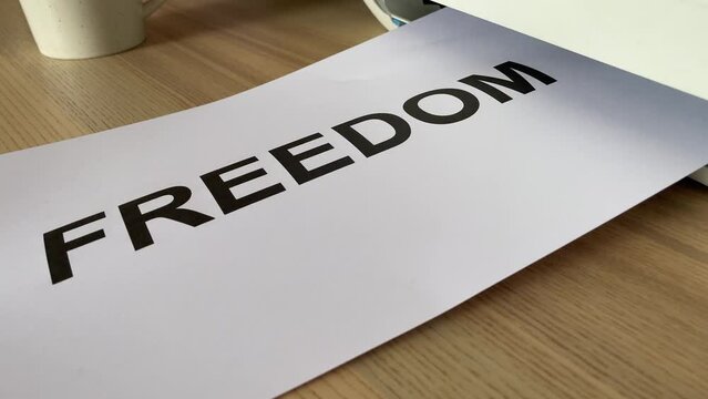 Printing &ldquo;FREEDOM&rdquo; text written message on paper sheet document. Close up A4 page being printed from multifunction scanner printer on wooden desk in office. Hand take paper from printer