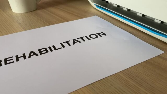 Printing &ldquo;REHABILITATION&rdquo; black text written message word on paper sheet A4 document. Close up page being printed from multifunction printer scanner in office. Hand take paper from printer