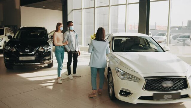 Professional saleswoman showing to multi ethnic couple various luxury cars at showroom. Happy african man and caucasian woman choosing new vehicle together. People wearing medical mask